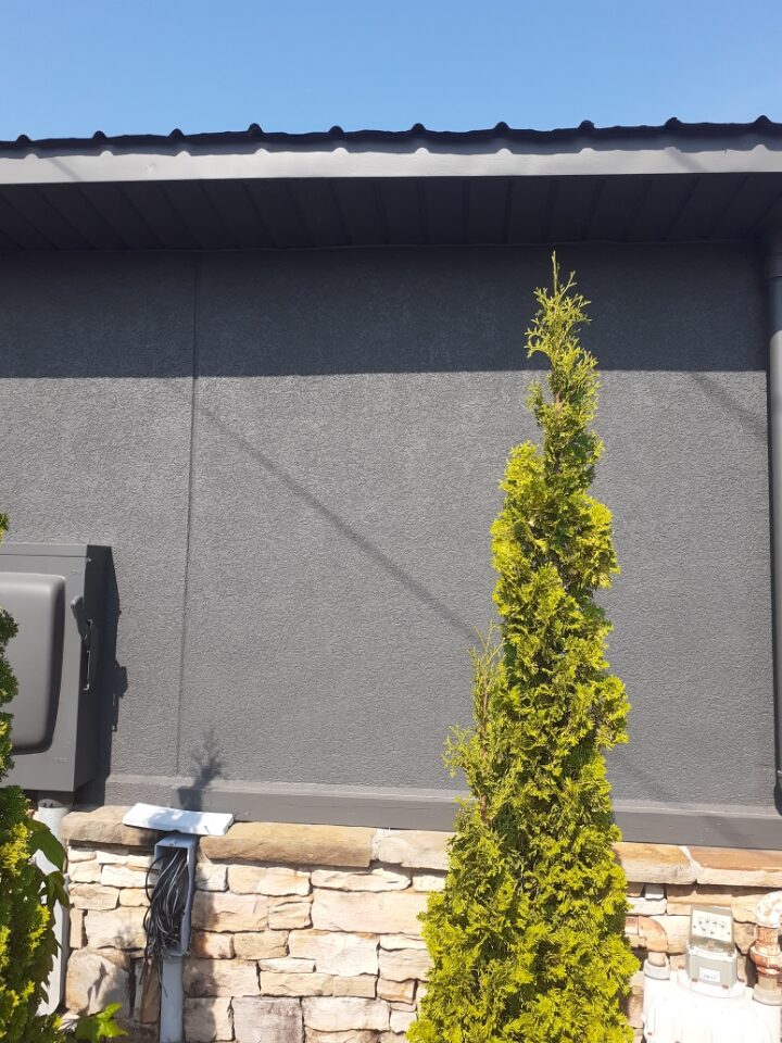 End wall of the same building in dark charcoal stucco running unbroken from a stone base up to the soffit