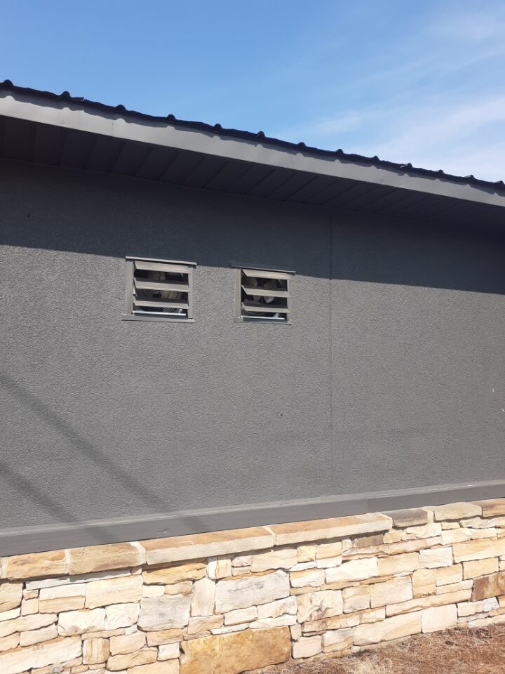 Side wall of the same building in dark charcoal stucco with two small louvred vents set into the wall above a stone base